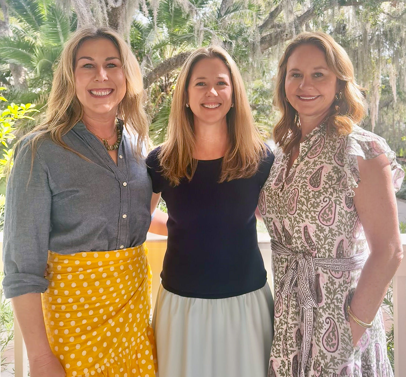 Three women standing together outdoors with trees and greenery in the background